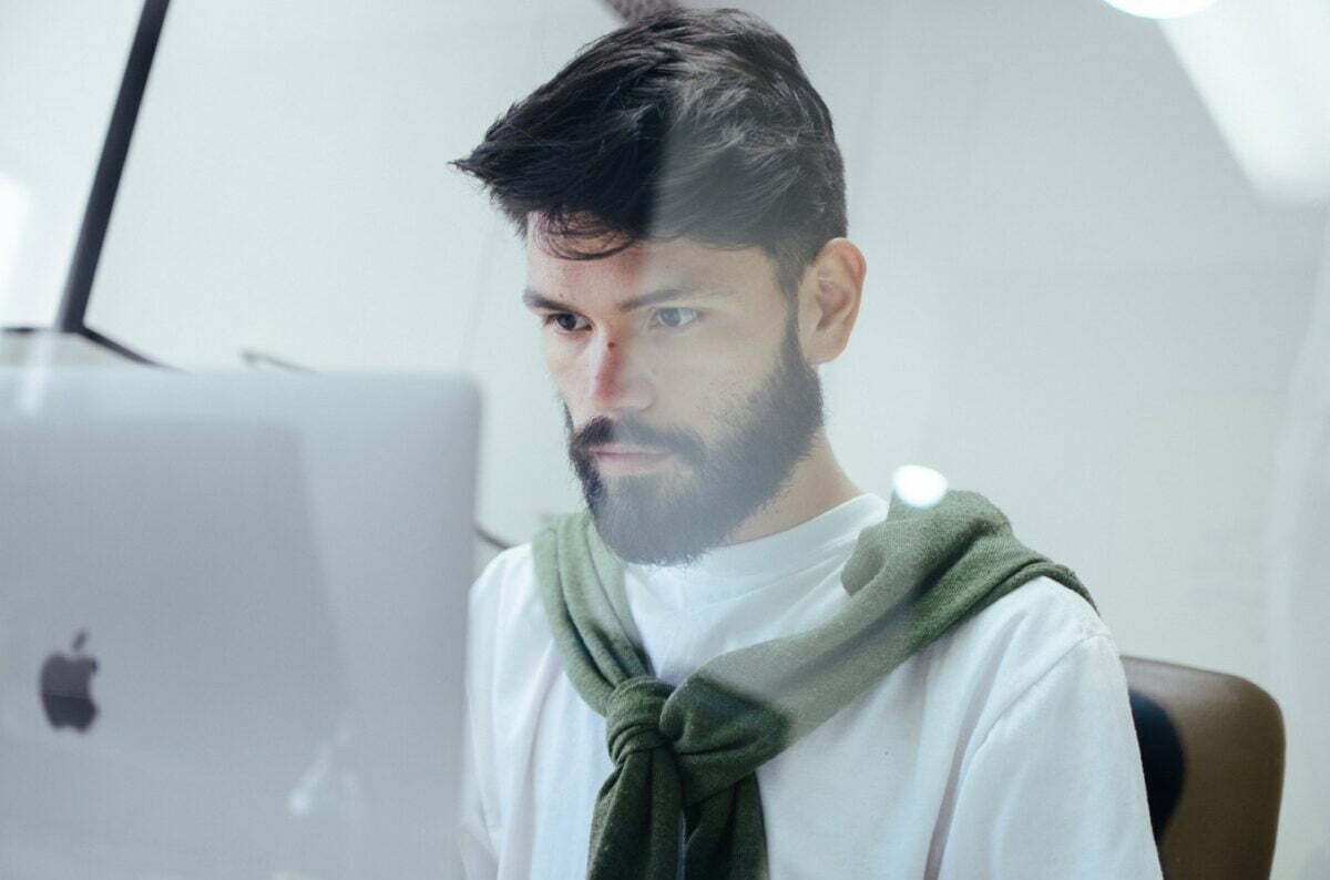 A person in a white shirt with a green sweater over his shoulders working with deep focus while looking at an Apple Screen