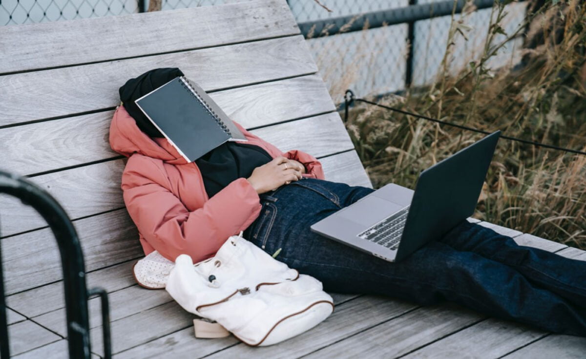 A person next to a white bag wearing a pink jacket. They have a laptop in their lap and are procrastinating, having a book covering their face.