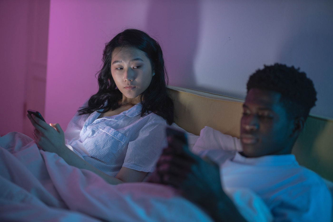 Two people sitting in purple-colored light and white shirts in bed looking at their phone