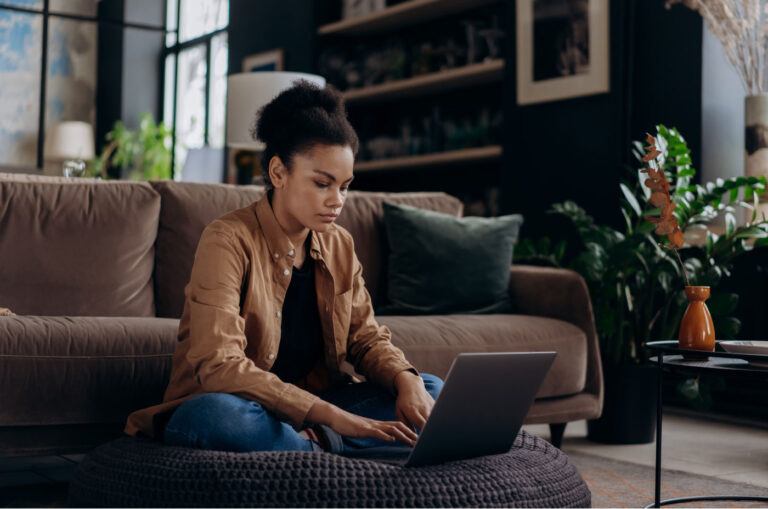 A person in a brown jacket sitting on the ground in front of their couch while on the laptop